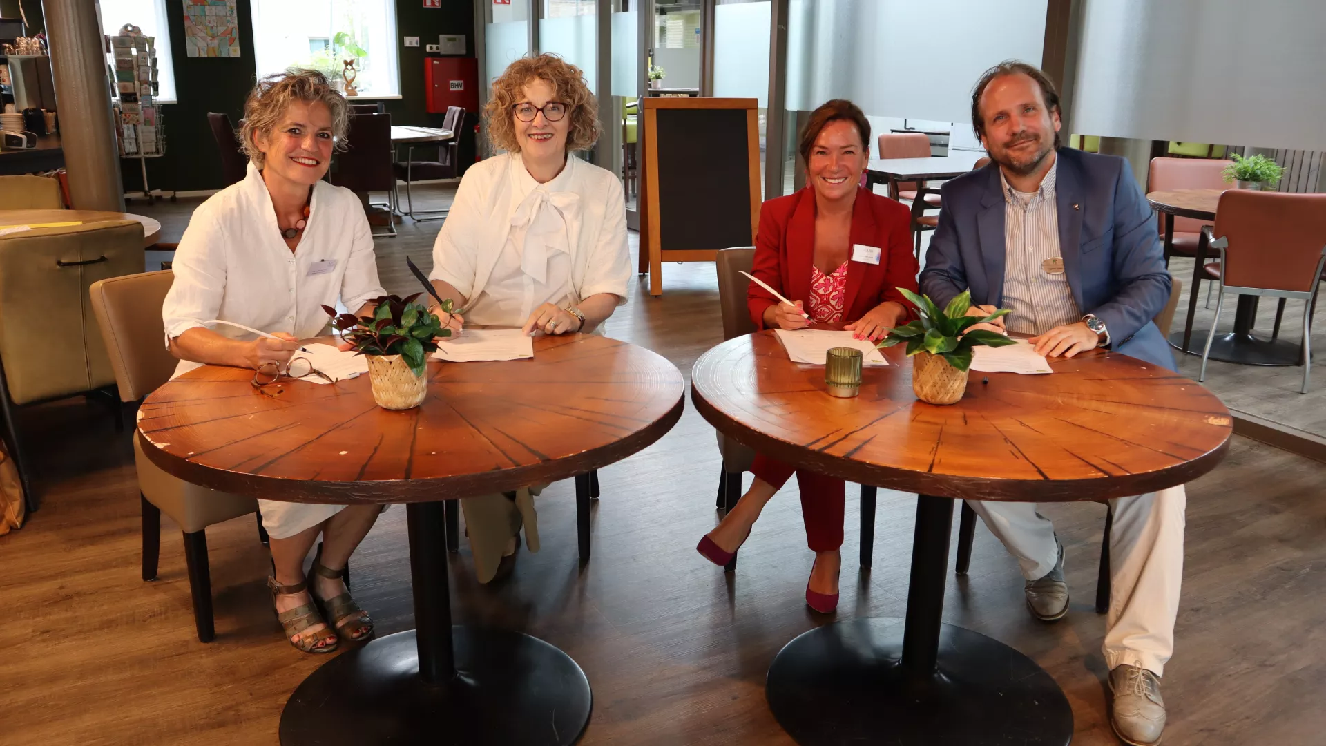 Bestuurders zitten aan ronde tafels en glimlachen naar de camera.